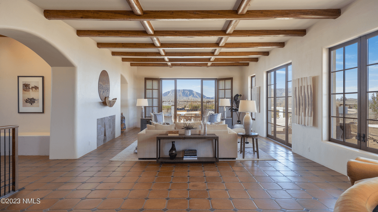 Interior of a staged Albuquerque home for sale, showing a bright living room with Saltillo tile floors, exposed vigas, and large windows with mountain views, professionally photographed for a real estate listing.