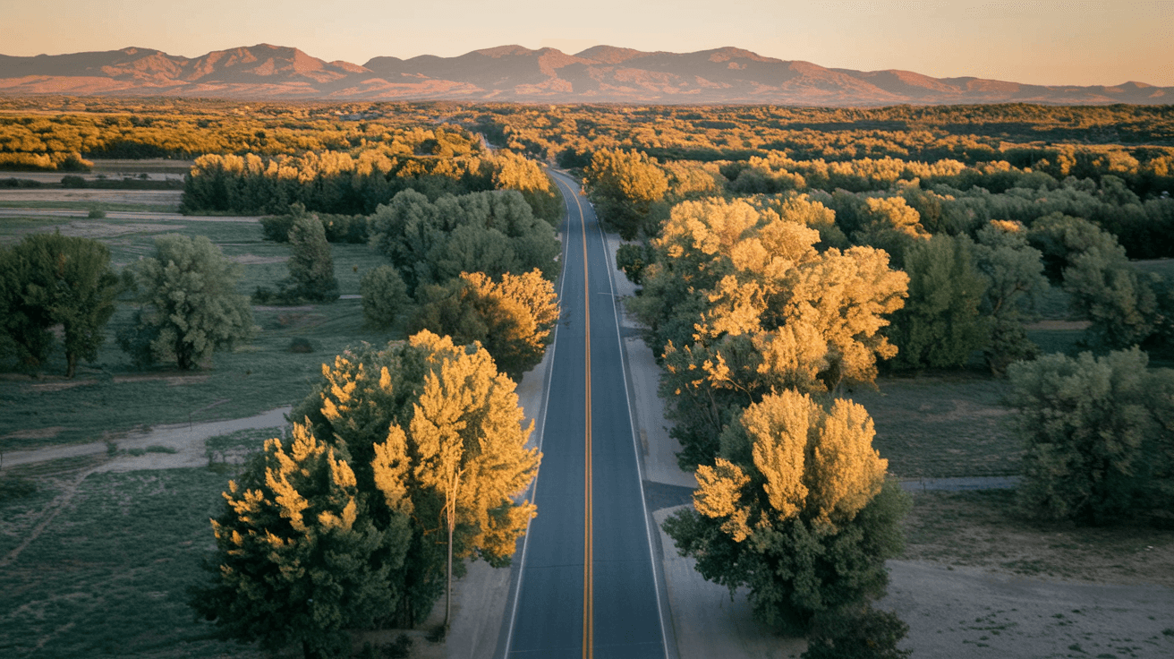 Aerial view of Corrales Road winding through green cottonwood bosque with the Sandia Mountains glowing at golden hour in the background