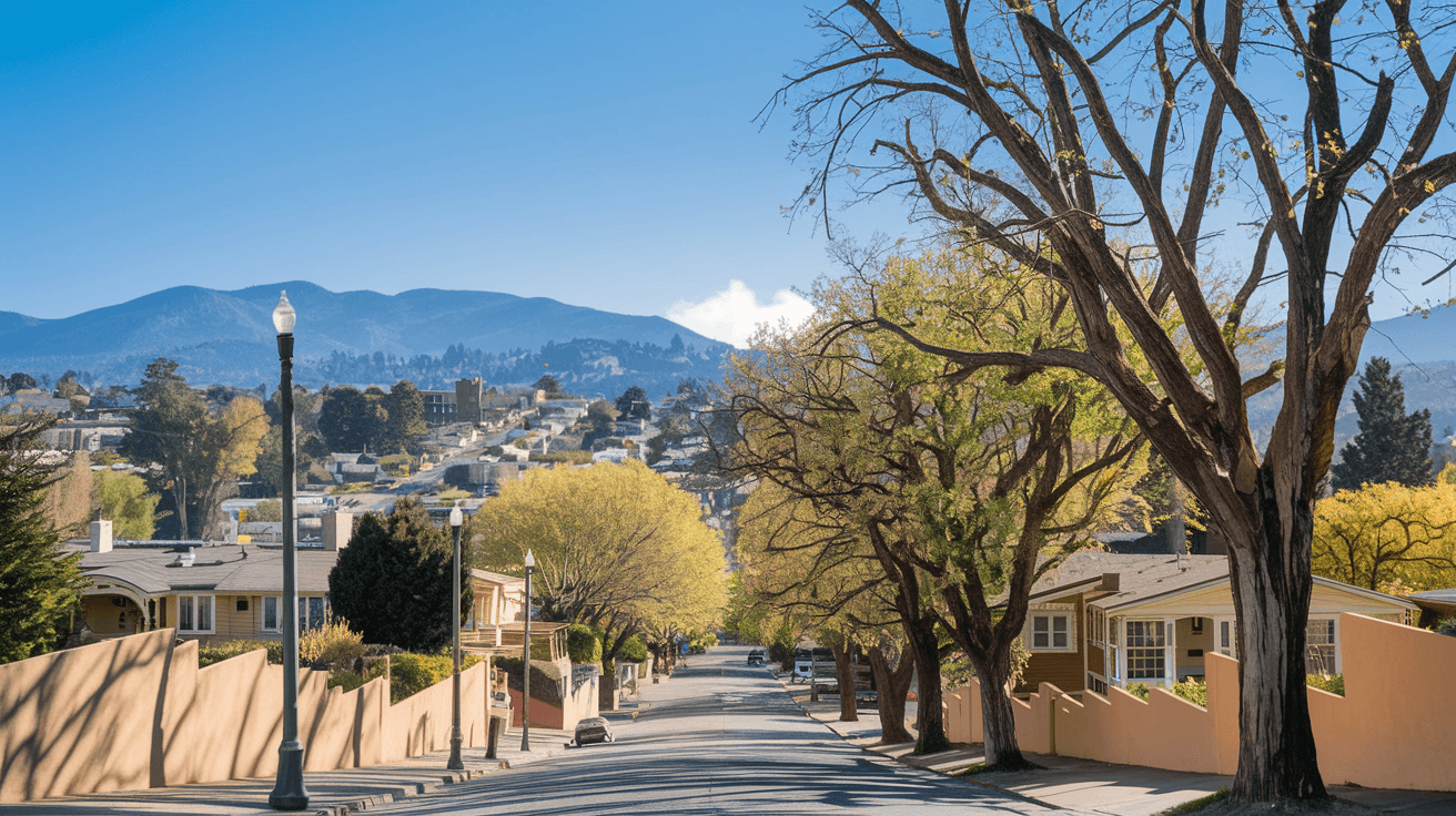 A tree-lined residential street in Nob Hill with mature cottonwoods, historic bungalows set back behind low adobe walls, and the Sandia Mountains visible in the background under a clear blue sky