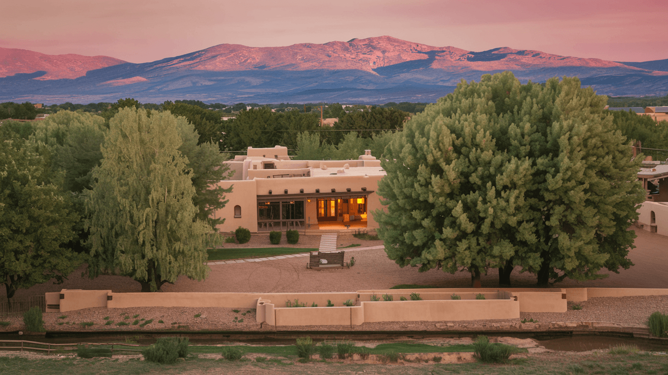 A sun-drenched historic adobe home on a wide North Valley lot with mature cottonwood trees, an acequia running along the property edge, and the Sandia Mountains glowing pink in the background at dusk