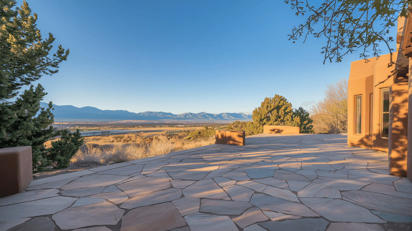 A wide flagstone patio on a Sandia Heights home overlooking the Rio Grande valley and West Mesa, with juniper trees framing the view and the late afternoon sun casting long shadows across the desert landscape