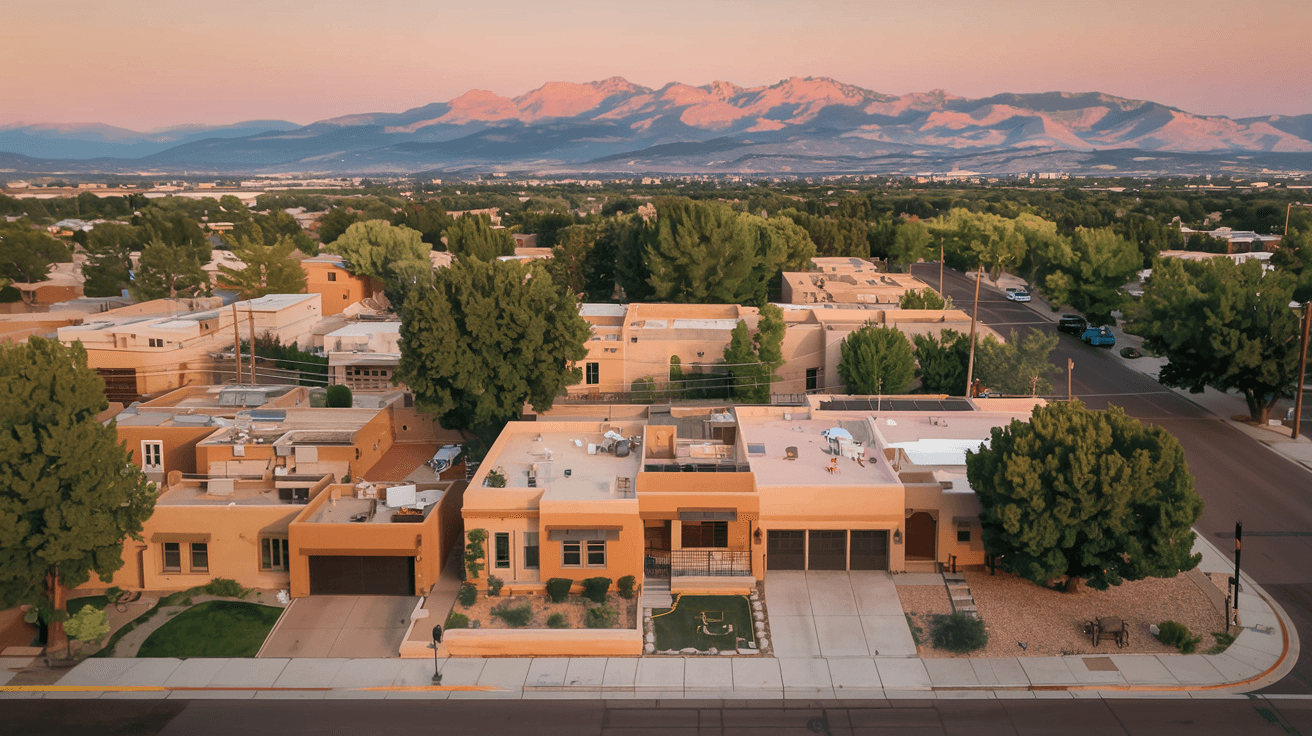 Aerial view of an Albuquerque neighborhood with adobe-style homes, the Sandia Mountains glowing pink at sunset in the background, and wide tree-lined streets below