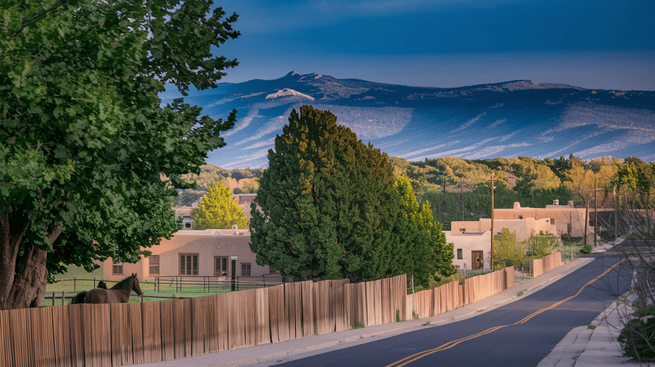 Tree-lined street in Corrales NM with historic adobe homes behind wooden fences, horses visible in a pasture, and the Sandia Mountains in the distant background under a deep blue sky